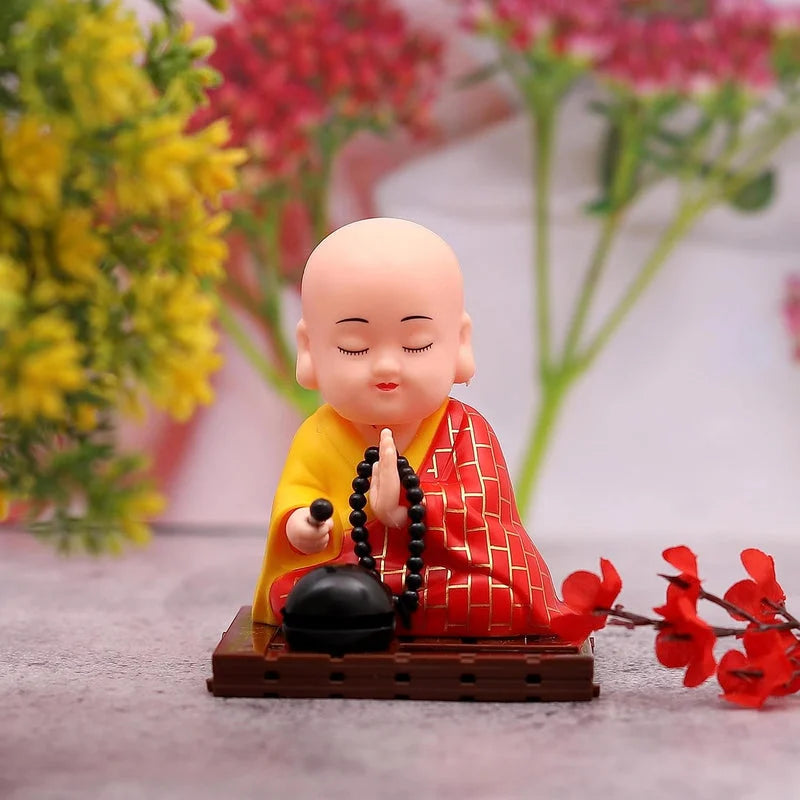 Small Buddha statue with bald head in red patterned robe, prayer beads in hands, on base