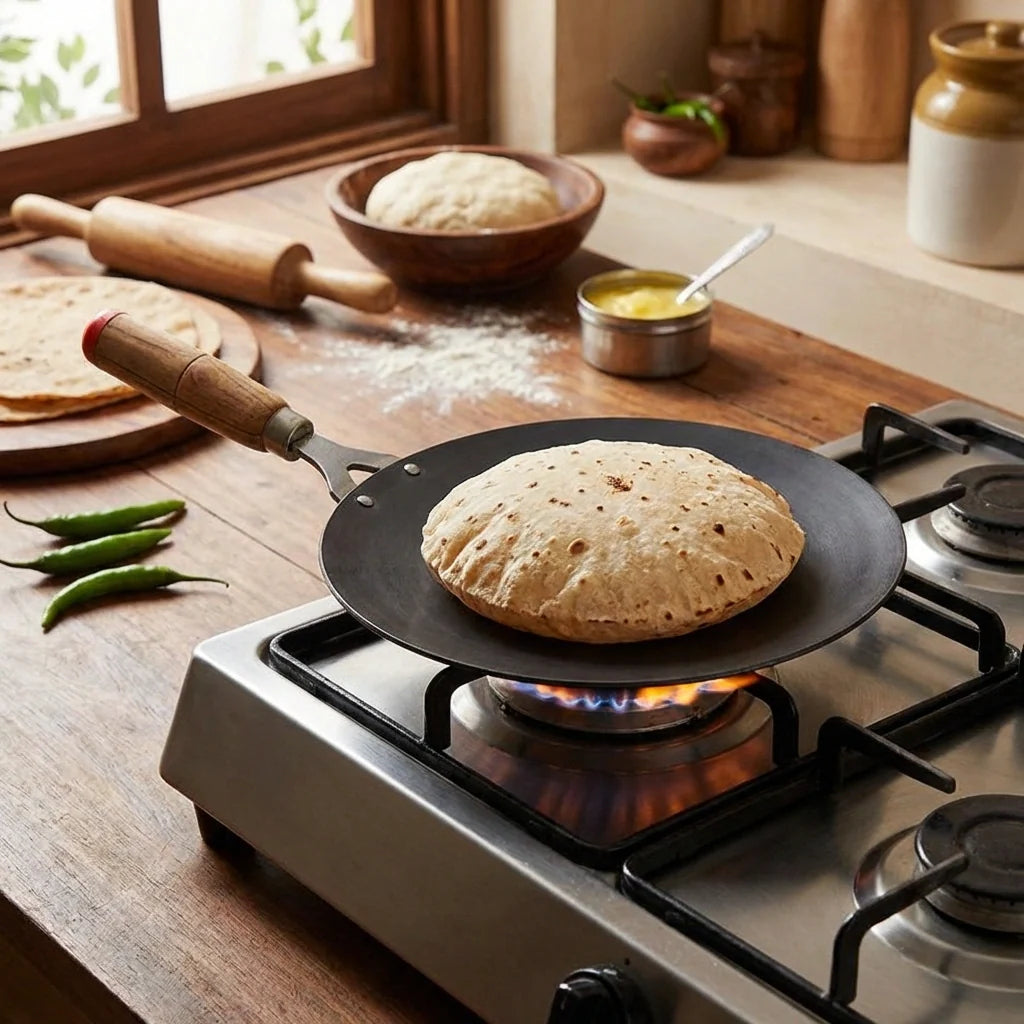 Wooden-handle roti tawa skillet on gas stove, roti cooking on black surface.
